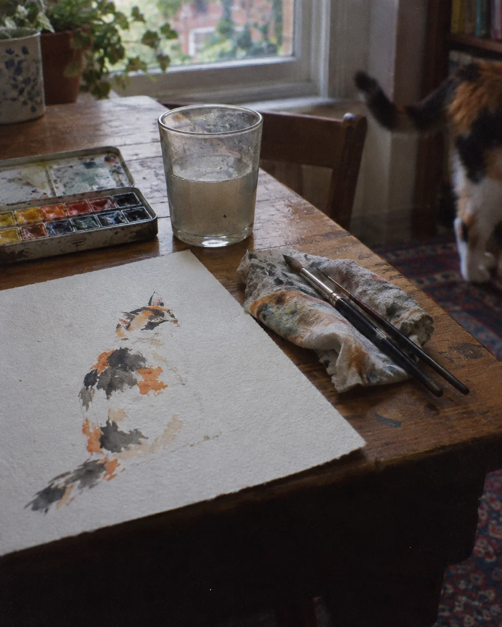 Freddie Mercury le declaró su amor a una gata en una canción de Queen y la acusó de orinar sus muebles 5 Boceto de acuarela sin terminar de una gata tricolor sobre papel texturizado, junto a una pequeña caja metálica de acuarelas, en una mesa de madera