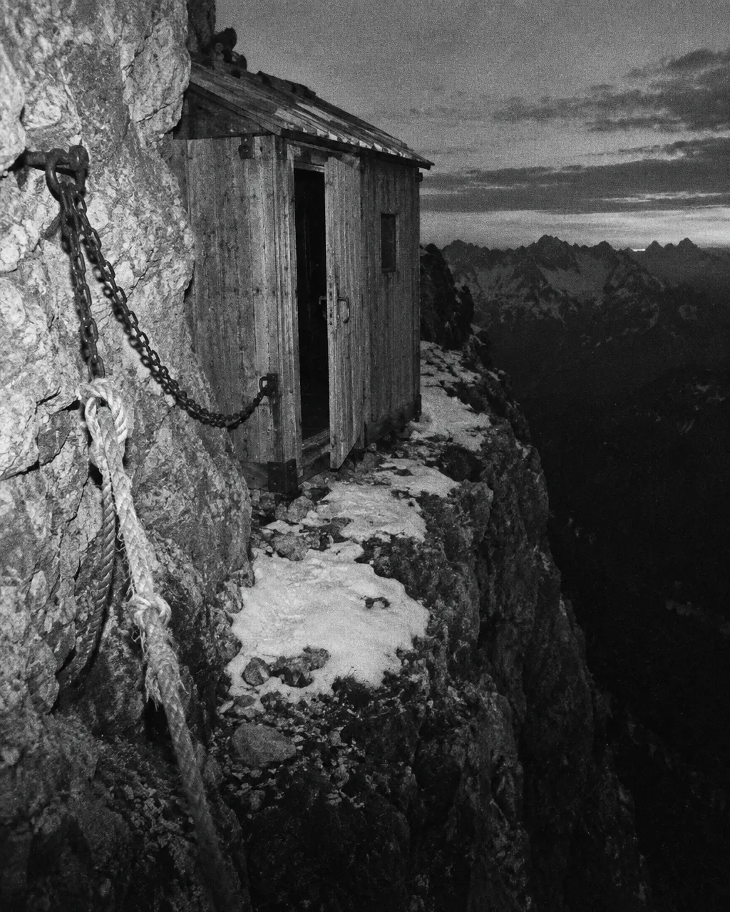 En 1950, un gatito salió detrás de los alpinistas del Cervino y siguió subiendo solo 3 Fotografía granulada en blanco y negro de una pequeña caseta de madera anclada a un saliente de roca en una arista empinada, al anochecer, con la puerta entreabierta, una cuerda y cadenas de hierro fijadas a la roca y nieve dura en el borde