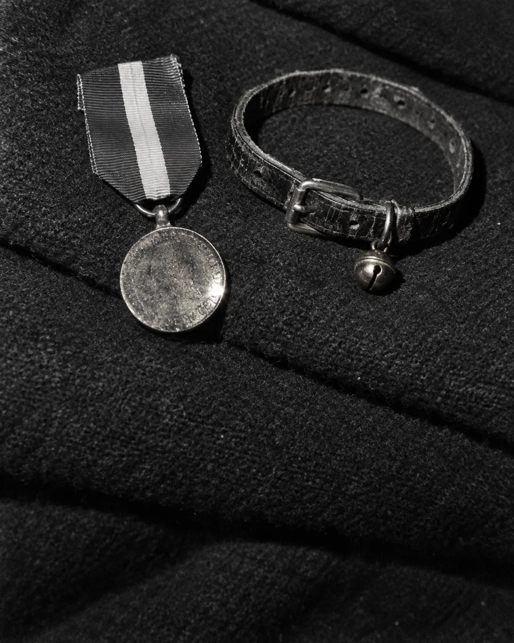 Medalla de bronce con cinta de tres franjas verticales junto a un collar de cuero gastado con un pequeño cascabel, sobre una manta naval oscura