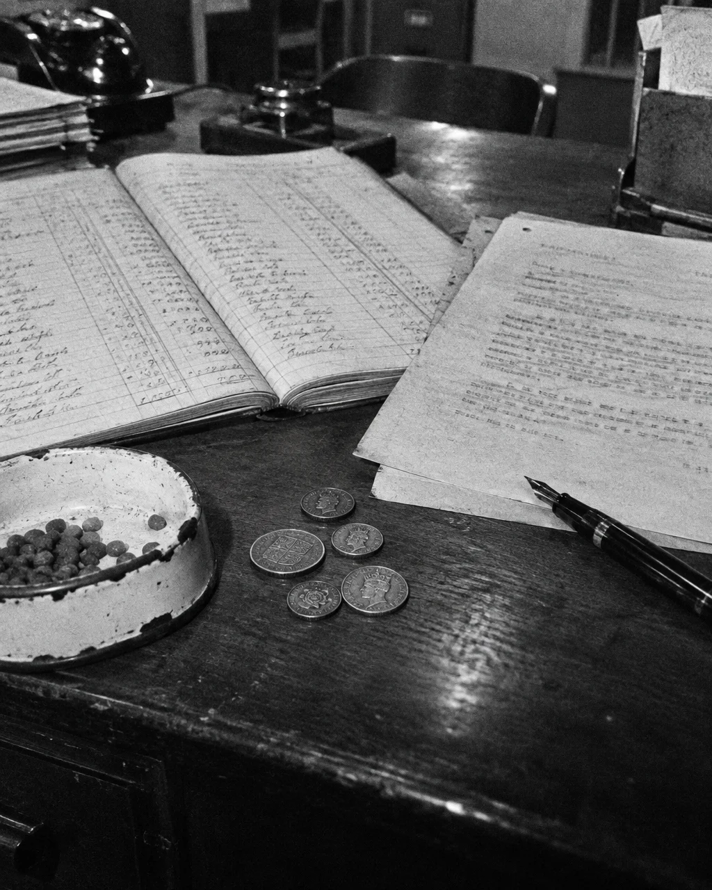 El correo británico les pagaba sueldo a sus gatos, y en 1953 el asunto llegó al Parlamento 4 Fotografía granulada en blanco y negro de un escritorio de madera en una oficina pública británica con un libro de cuentas abierto con columnas manuscritas, un memorando mecanografiado, varias monedas británicas antiguas y gastadas, un pequeño plato de esmalte descascarado para gato y una pluma estilográfica.