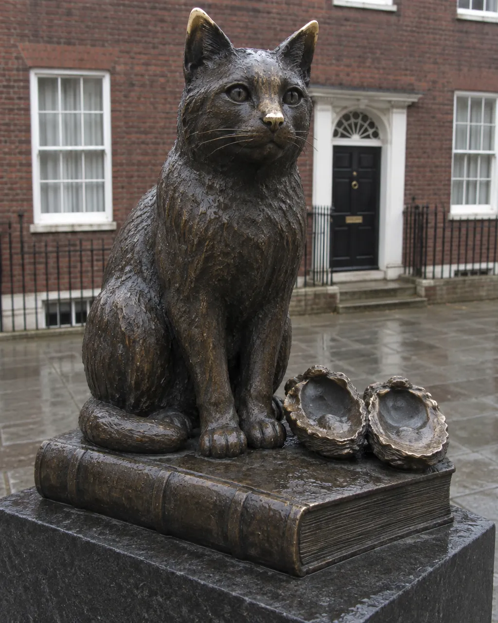 Gato de bronce sentado sobre un gran libro cerrado, también de bronce, en un patio pavimentado bajo un cielo nublado