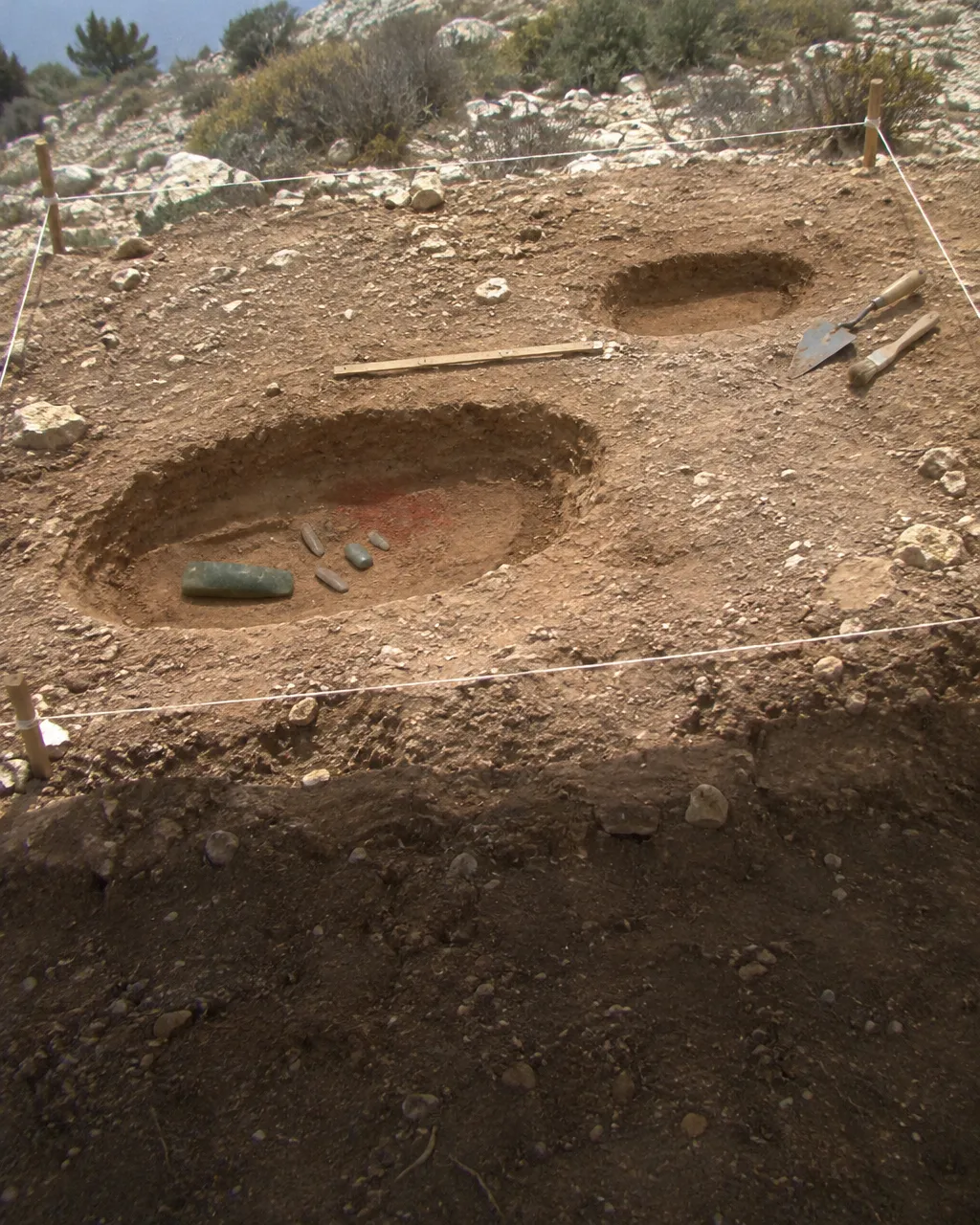 Excavación neolítica en una ladera de caliza con matorral seco, vista desde arriba, con una cuadrícula de cuerdas tendida sobre tierra clara y rojiza