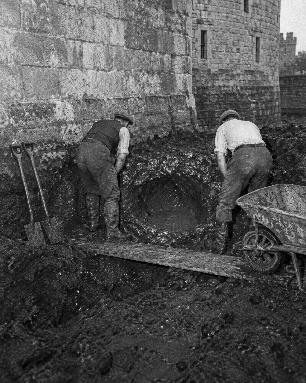 Zanja profunda abierta en arcilla húmeda y oscura al pie de una muralla de piedra gris, en blanco y negro con grano de película