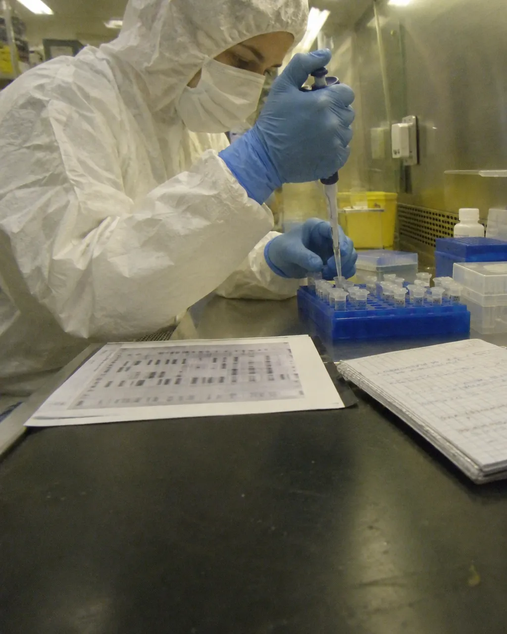 Persona con traje blanco completo, capucha, mascarilla y guantes azules trabajando en un laboratorio pequeño y limpio
