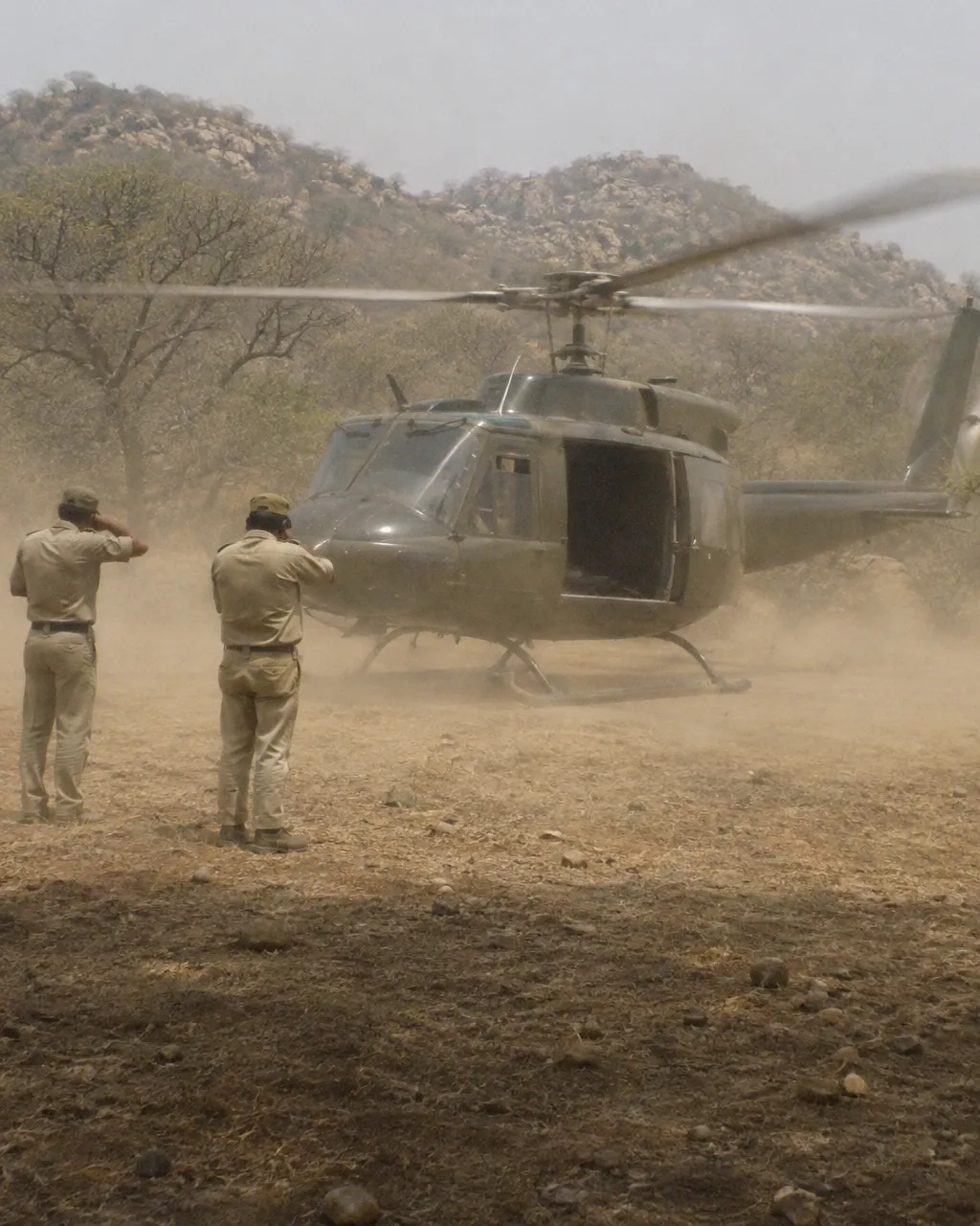 Un helicóptero recién posado en un claro polvoriento de un bosque seco, con las aspas girando y dos guardabosques de uniforme caqui cubriéndose la cara del polvo
